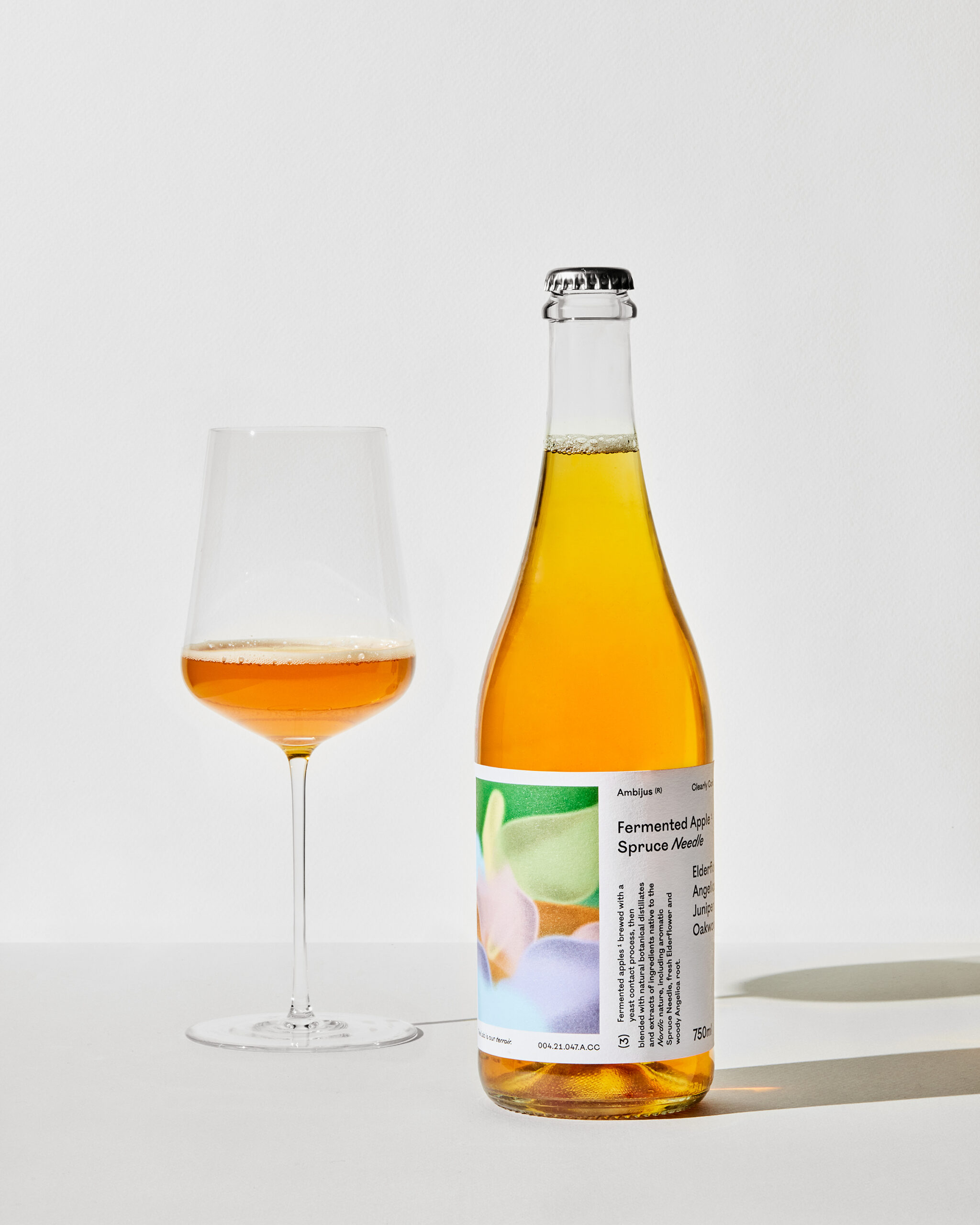 Flaske og vinglass med Ambijus 3: Clearly Confused – en alkoholfri, fermentert drikk laget med epler fra Hardanger, granbar, hylleblomst, kvannrot, einebær og eik.