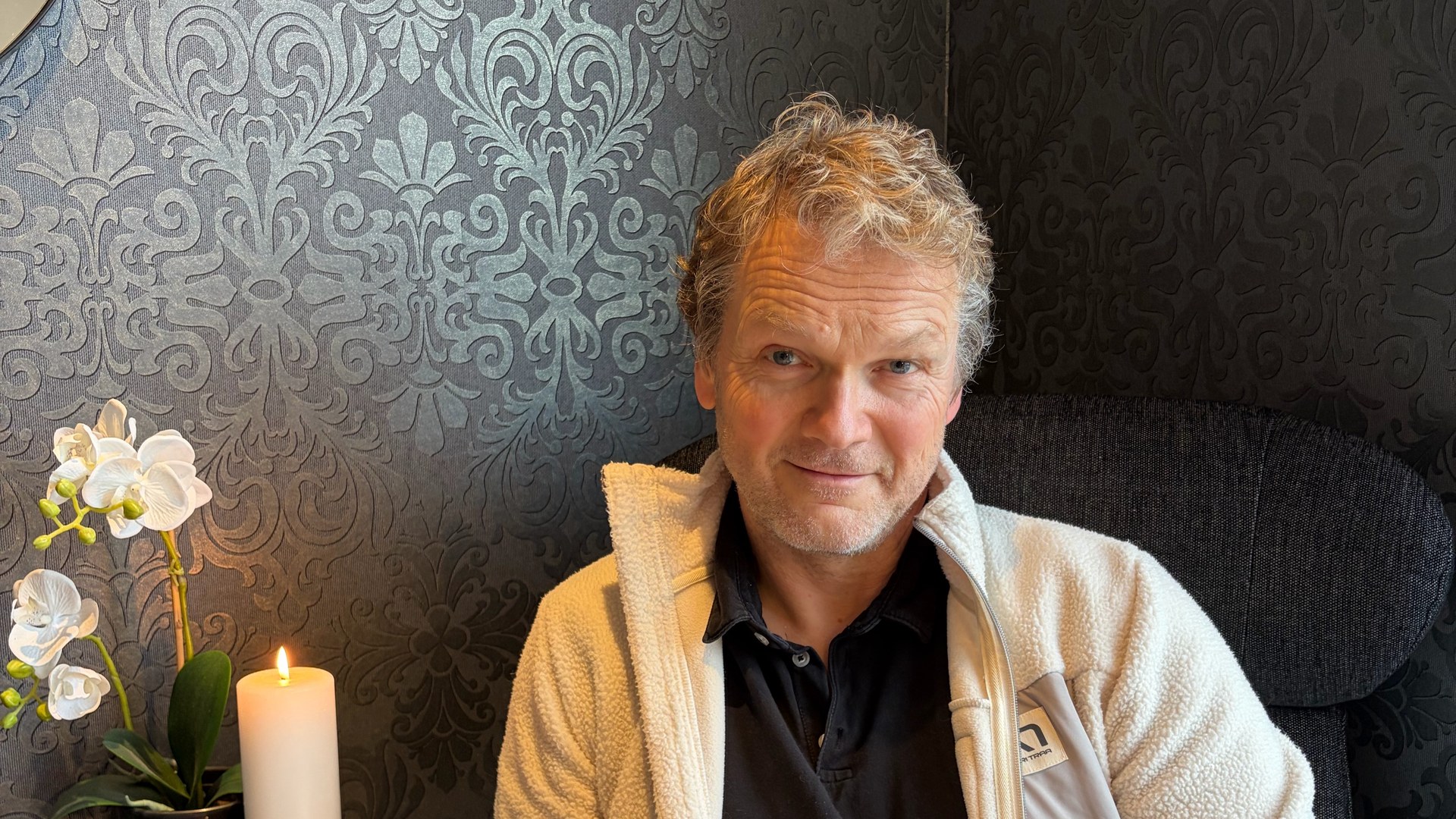 Henrik Berntsen sitter i en stol med en kopp te, foran en mønstret tapet med et tent stearinlys og en orkidé.